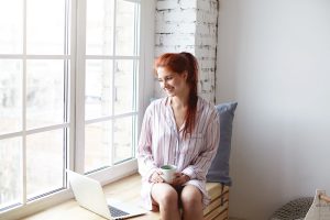 beautiful-young-female-with-cute-smile-ginger-hair-ponytail-enjoying-slow-morning-home-sitting-by-large-window-drinking-coffee-surfing-internet-laptop-daylight-sunshine-soft-colors